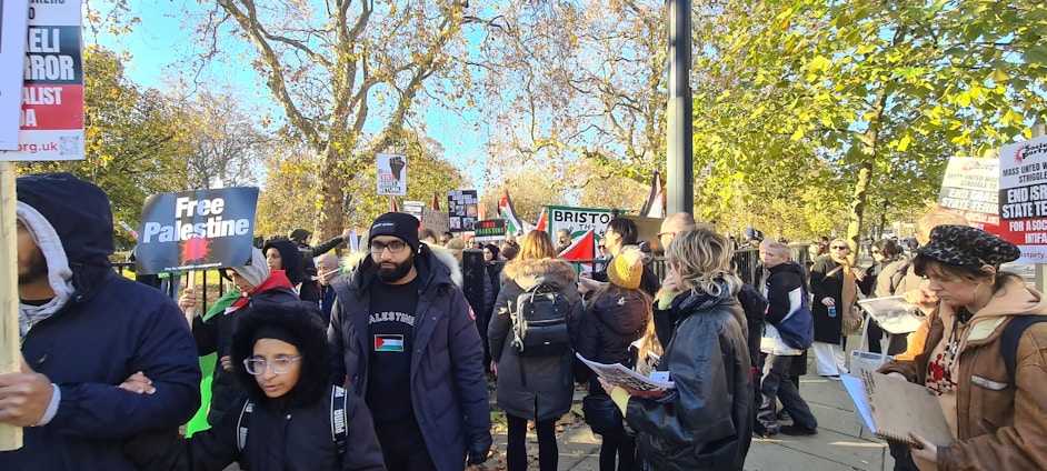 A group of people is gathered outdoors, holding signs and banners with messages related to political activism. The scene takes place on a sunny day with trees in the background showing autumn colors. Many people are wearing jackets, indicating cool weather.