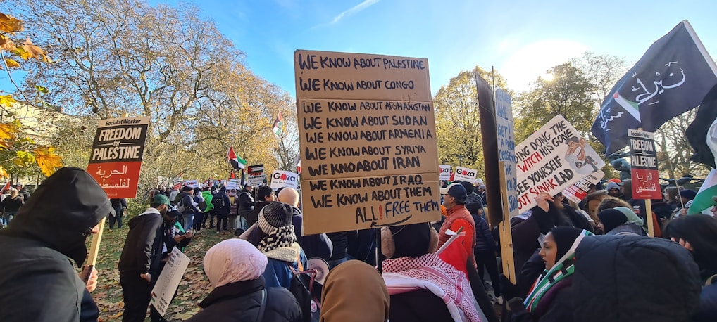 A large group of people gathered outdoors, many holding signs with messages about global political issues and freedom for various countries. The crowd appears diverse, with individuals wearing various types of clothing suitable for cooler weather. Several trees with autumn leaves are in the background, suggesting the season is fall.