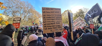 A large group of people gathered outdoors, many holding signs with messages about global political issues and freedom for various countries. The crowd appears diverse, with individuals wearing various types of clothing suitable for cooler weather. Several trees with autumn leaves are in the background, suggesting the season is fall.