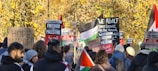 A group of people gathered in a protest, holding signs and banners with messages related to freedom for Palestine and social justice. Some of the signs read 'HOPE MARCH,' 'Forward to Palestine,' and 'End Israeli State Terror.' The crowd is diverse and features an assortment of ethnic backgrounds. In the background, there are trees with autumn-colored leaves, adding a natural backdrop to the demonstration.