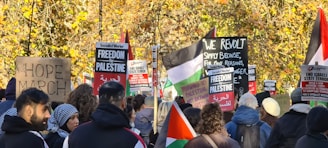 A group of people gathered in a protest, holding signs and banners with messages related to freedom for Palestine and social justice. Some of the signs read 'HOPE MARCH,' 'Forward to Palestine,' and 'End Israeli State Terror.' The crowd is diverse and features an assortment of ethnic backgrounds. In the background, there are trees with autumn-colored leaves, adding a natural backdrop to the demonstration.
