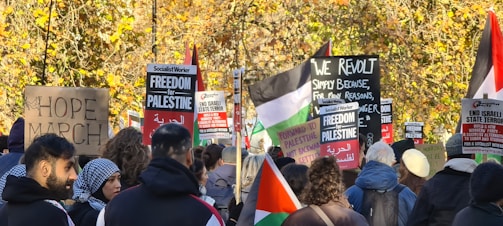 A group of people gathered in a protest, holding signs and banners with messages related to freedom for Palestine and social justice. Some of the signs read 'HOPE MARCH,' 'Forward to Palestine,' and 'End Israeli State Terror.' The crowd is diverse and features an assortment of ethnic backgrounds. In the background, there are trees with autumn-colored leaves, adding a natural backdrop to the demonstration.