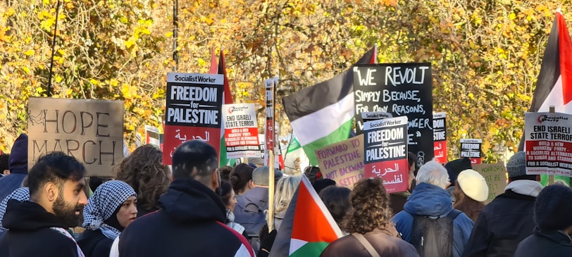 A group of people gathered in a protest, holding signs and banners with messages related to freedom for Palestine and social justice. Some of the signs read 'HOPE MARCH,' 'Forward to Palestine,' and 'End Israeli State Terror.' The crowd is diverse and features an assortment of ethnic backgrounds. In the background, there are trees with autumn-colored leaves, adding a natural backdrop to the demonstration.