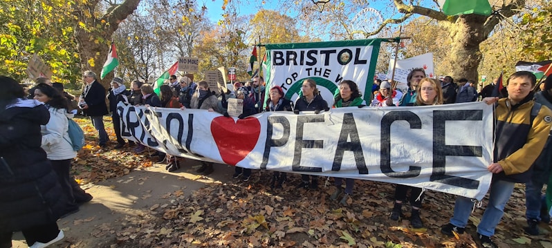 A group of people holding banners in an outdoor setting with autumn leaves on the ground. The banners have messages advocating for peace and stopping war, with flags in the background.