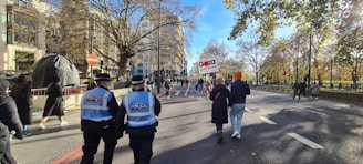 A street scene featuring a group of people walking along a broad road lined with trees, with some holding signs. Two police officers with high-visibility vests are visible on the left side. The road is free of vehicles and bordered by trees with autumn foliage, and a few buildings are visible in the background.