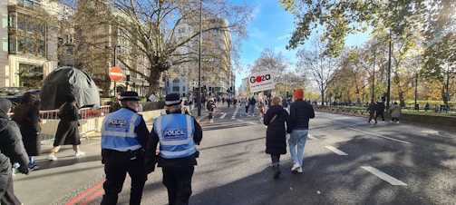 A street scene featuring a group of people walking along a broad road lined with trees, with some holding signs. Two police officers with high-visibility vests are visible on the left side. The road is free of vehicles and bordered by trees with autumn foliage, and a few buildings are visible in the background.