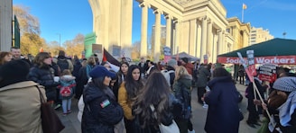 A large group of people are gathered in front of a neoclassical structure holding various signs. Among the banners and signs, many read 'Freedom for Palestine' and 'Socialist Workers'. The weather appears to be clear and sunny. The event is taking place in what seems to be an urban park or public space with trees and modern buildings in the background.