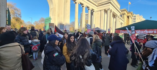 A large group of people are gathered in front of a neoclassical structure holding various signs. Among the banners and signs, many read 'Freedom for Palestine' and 'Socialist Workers'. The weather appears to be clear and sunny. The event is taking place in what seems to be an urban park or public space with trees and modern buildings in the background.