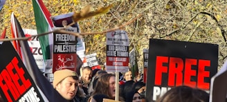 A group of people gathered outdoors, holding signs with messages advocating for freedom and protesting against Israeli state actions. The signs have bold red and black text with phrases like 'FREEDOM for PALESTINE' and 'END ISRAELI STATE TERROR'. The crowd appears engaged and passionate, framed by autumn trees with golden leaves.