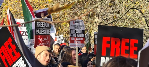 A group of people gathered outdoors, holding signs with messages advocating for freedom and protesting against Israeli state actions. The signs have bold red and black text with phrases like 'FREEDOM for PALESTINE' and 'END ISRAELI STATE TERROR'. The crowd appears engaged and passionate, framed by autumn trees with golden leaves.