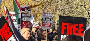 A group of people gathered outdoors, holding signs with messages advocating for freedom and protesting against Israeli state actions. The signs have bold red and black text with phrases like 'FREEDOM for PALESTINE' and 'END ISRAELI STATE TERROR'. The crowd appears engaged and passionate, framed by autumn trees with golden leaves.