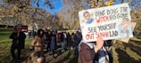 A group of people are participating in a protest march in a park. One person is holding a sign with political slogans and caricatures, while another sign reads 'Freedom for Palestine'. The scene is set under a clear blue sky with trees displaying autumn foliage.