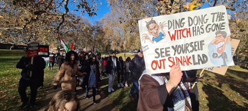 A group of people are participating in a protest march in a park. One person is holding a sign with political slogans and caricatures, while another sign reads 'Freedom for Palestine'. The scene is set under a clear blue sky with trees displaying autumn foliage.
