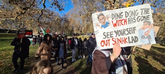 A group of people are participating in a protest march in a park. One person is holding a sign with political slogans and caricatures, while another sign reads 'Freedom for Palestine'. The scene is set under a clear blue sky with trees displaying autumn foliage.