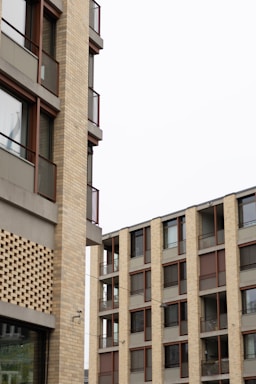 A modern architectural structure featuring multiple multi-story buildings. The exteriors are composed of brick and concrete with large windows and metal railings. The design is geometric, with clean lines and grids formed by the windows and balconies.