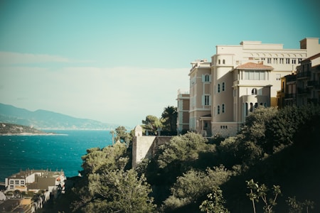 A coastal scene features a large, elegant building with an architectural style reminiscent of Mediterranean villas, situated on a cliffside. The structure is surrounded by lush greenery, and below is a view of the blue sea. Distant mountains are visible under a clear sky, enhancing the picturesque landscape.