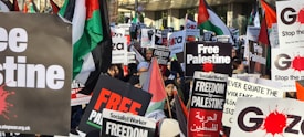 A large group of people demonstrating with banners and placards advocating for Palestine. The signs are prominently displaying slogans like 'Free Palestine' and have a mix of English and Arabic text. Several Palestinian flags are visible among the crowd.
