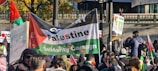 A crowd of people gathered in an outdoor setting, holding flags and signs supporting a Palestinian solidarity campaign. The main banner prominently displays colors associated with Palestine—red, black, and green—along with text in support of Palestine. The surroundings include a fence and some trees with autumn foliage.