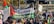 A crowd of people gathered in an outdoor setting, holding flags and signs supporting a Palestinian solidarity campaign. The main banner prominently displays colors associated with Palestine—red, black, and green—along with text in support of Palestine. The surroundings include a fence and some trees with autumn foliage.