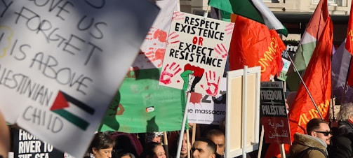 A vibrant protest scene with a crowd holding various signs and flags. Prominently featured are signs with messages supporting Palestinian rights, including 'Respect Existence or Expect Resistance'. Red handprints and a clenched fist graphic are visible on one sign. Several large red, green, and black flags are waving in the background, amidst the bustling crowd.
