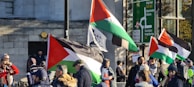 A group of people are gathered in a public space holding Palestinian flags. The setting is urban, with a stone building and street signs visible. Some individuals are engaged in conversation while others are observing the surroundings. The crowd includes men and women dressed warmly, suggesting cool weather.