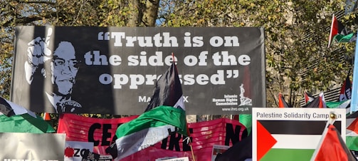 A protest scene with a large banner featuring a portrait and the phrase 'Truth is on the side of the oppressed' attributed to Malcolm X. Numerous flags, including Palestinian flags, and another banner for the Palestine Solidarity Campaign are visible. The setting appears to be outdoors with trees in the background.