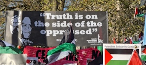 A protest scene with a large banner featuring a portrait and the phrase 'Truth is on the side of the oppressed' attributed to Malcolm X. Numerous flags, including Palestinian flags, and another banner for the Palestine Solidarity Campaign are visible. The setting appears to be outdoors with trees in the background.