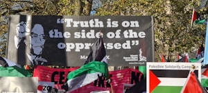A protest scene with a large banner featuring a portrait and the phrase 'Truth is on the side of the oppressed' attributed to Malcolm X. Numerous flags, including Palestinian flags, and another banner for the Palestine Solidarity Campaign are visible. The setting appears to be outdoors with trees in the background.