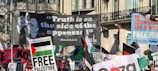 A crowded protest scene featuring numerous banners and flags. The prominent large banner displays a quote attributed to Malcolm X, reading 'Truth is on the side of the oppressed'. Other visible signs include messages supporting Palestine and opposing Zionism and Islamophobia.