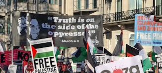 A crowded protest scene featuring numerous banners and flags. The prominent large banner displays a quote attributed to Malcolm X, reading 'Truth is on the side of the oppressed'. Other visible signs include messages supporting Palestine and opposing Zionism and Islamophobia.
