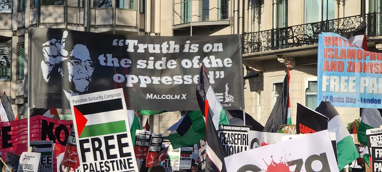 A crowded protest scene featuring numerous banners and flags. The prominent large banner displays a quote attributed to Malcolm X, reading 'Truth is on the side of the oppressed'. Other visible signs include messages supporting Palestine and opposing Zionism and Islamophobia.
