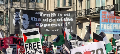 A crowded protest scene featuring numerous banners and flags. The prominent large banner displays a quote attributed to Malcolm X, reading 'Truth is on the side of the oppressed'. Other visible signs include messages supporting Palestine and opposing Zionism and Islamophobia.