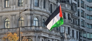A large flag with red, white, black, and green colors is prominently displayed against the backdrop of an ornate historical building. The architecture features intricate designs with arched windows and decorative columns. Alongside, a modern building with geometric patterns contrasts with the traditional architecture. Some autumn leaves are visible in a tree in the foreground.