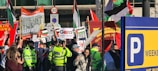 A crowded street protest featuring diverse groups of people holding various signs and banners related to political and social issues. Police officers in bright yellow jackets observe the gathering. Flags, including Palestinian flags, and signs with messages of solidarity and calls for freedom are prominent. There is a mix of color and activity against an urban backdrop, indicating a vibrant and engaged scene.