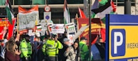 A crowded street protest featuring diverse groups of people holding various signs and banners related to political and social issues. Police officers in bright yellow jackets observe the gathering. Flags, including Palestinian flags, and signs with messages of solidarity and calls for freedom are prominent. There is a mix of color and activity against an urban backdrop, indicating a vibrant and engaged scene.