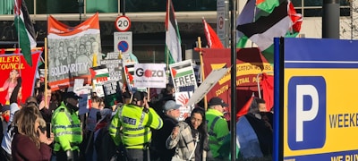 A crowded street protest featuring diverse groups of people holding various signs and banners related to political and social issues. Police officers in bright yellow jackets observe the gathering. Flags, including Palestinian flags, and signs with messages of solidarity and calls for freedom are prominent. There is a mix of color and activity against an urban backdrop, indicating a vibrant and engaged scene.