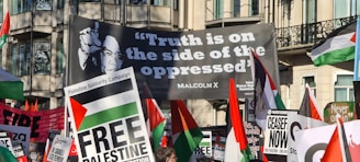 A protest scene with numerous signs and flags advocating for Palestinian solidarity. The prominent banner carries a quote from Malcolm X, 'Truth is on the side of the oppressed,' accompanied by his image, demonstrating a message of support and unity. The crowd holds various placards with slogans such as 'Free Palestine' and 'Ceasefire Now.' The scene is set in an urban environment with buildings in the background.