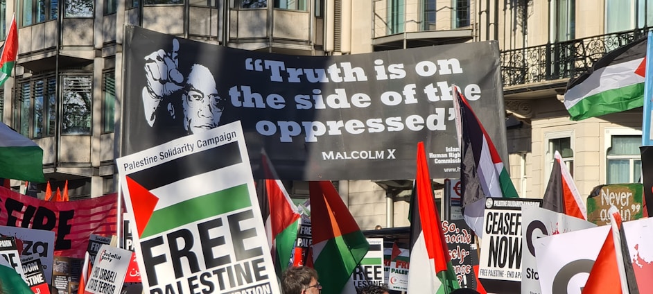 A protest scene with numerous signs and flags advocating for Palestinian solidarity. The prominent banner carries a quote from Malcolm X, 'Truth is on the side of the oppressed,' accompanied by his image, demonstrating a message of support and unity. The crowd holds various placards with slogans such as 'Free Palestine' and 'Ceasefire Now.' The scene is set in an urban environment with buildings in the background.