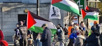 A group of people are gathered outdoors, some holding flags with a distinct red, black, white, and green color pattern. One flag has a silhouette of a person with text. Many individuals are wearing winter clothing, such as jackets and hats. There are also some signs and banners, with one that reads 'Free Palestine.' Some people appear to be having conversations or interacting with each other.