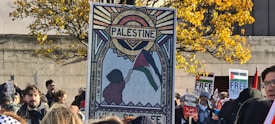 A group of people gathered outdoors, holding signs related to Palestine. A prominent sign features artwork depicting a person holding a flag with the sun and the word 'Palestine' above. A tree with yellow leaves and a stone wall are visible in the background.