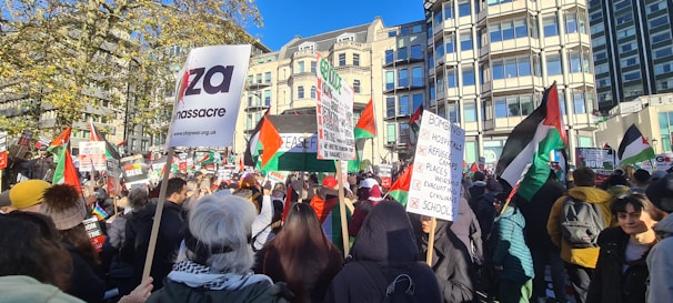 A large group of people gathered in a city square holding various signs and flags. Many flags have red, white, green, and black colors. The background features tall buildings and some trees under a clear blue sky. The crowd appears to be participating in a demonstration or protest, with messages regarding political and humanitarian issues visible on the signs.