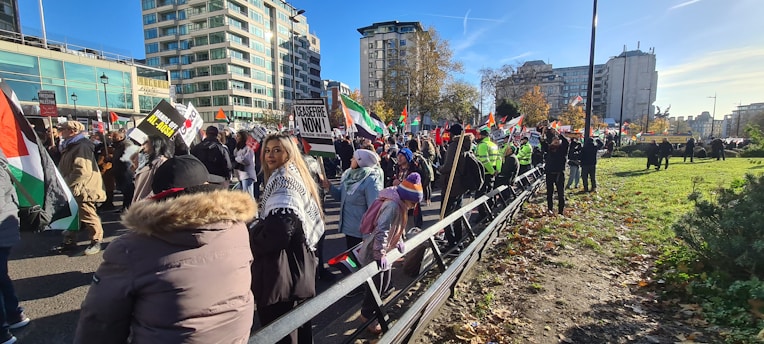 A crowd of people is gathered at a demonstration, holding signs and flags. The event takes place in an urban environment, surrounded by tall buildings under a clear blue sky. Many participants are dressed in winter clothing, and there is a noticeable presence of police in bright safety jackets.