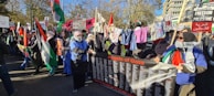 A large group of people is gathered in an outdoor space, holding various signs and banners related to Palestine. They are showing support through banners with messages like 'Angels of Gaza,' and waving Palestinian flags. Clothes are hung on a line, and many participants wear headscarves.