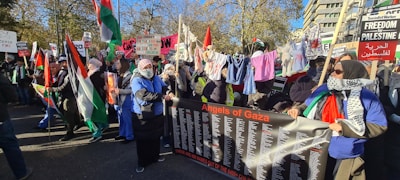 A large group of people is gathered in an outdoor space, holding various signs and banners related to Palestine. They are showing support through banners with messages like 'Angels of Gaza,' and waving Palestinian flags. Clothes are hung on a line, and many participants wear headscarves.