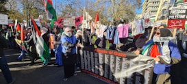 A large group of people is gathered in an outdoor space, holding various signs and banners related to Palestine. They are showing support through banners with messages like 'Angels of Gaza,' and waving Palestinian flags. Clothes are hung on a line, and many participants wear headscarves.