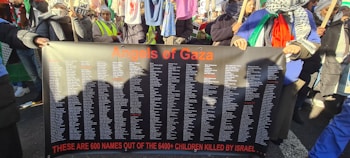 A group of people hold a large black banner with orange text reading 'Angels of Gaza' at the top. The banner lists over 600 names, with a message below stating these are some of the children killed in a conflict. The individuals are wearing scarves and holding flags, suggesting participation in a protest or demonstration. Clothing and accessories include patterned scarves and bright colored garments.