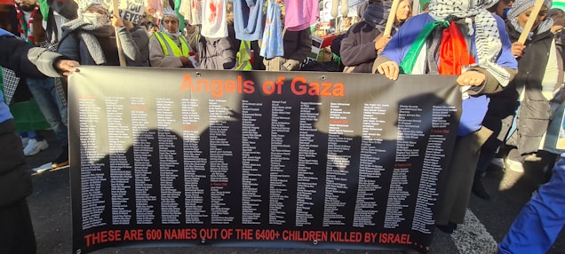 A group of people hold a large black banner with orange text reading 'Angels of Gaza' at the top. The banner lists over 600 names, with a message below stating these are some of the children killed in a conflict. The individuals are wearing scarves and holding flags, suggesting participation in a protest or demonstration. Clothing and accessories include patterned scarves and bright colored garments.