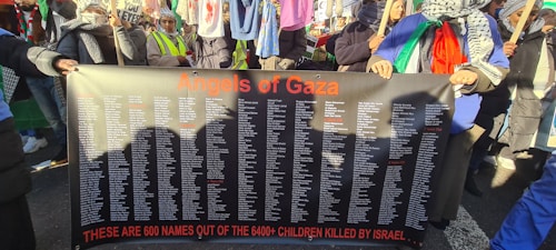 A group of people hold a large black banner with orange text reading 'Angels of Gaza' at the top. The banner lists over 600 names, with a message below stating these are some of the children killed in a conflict. The individuals are wearing scarves and holding flags, suggesting participation in a protest or demonstration. Clothing and accessories include patterned scarves and bright colored garments.