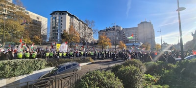 A large gathering of people in an urban area, featuring many individuals holding flags and banners. Several buildings surround the scene, and there are leafy green plants and trees. Police officers in high-visibility jackets are managing the crowd.