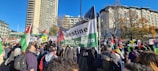 A large group of people gathered outdoors holding flags and signs. There are buildings and trees in the background, with a clear blue sky overhead. The signs and flags prominently feature the colors red, green, black, and white, and some have text related to a campaign or cause.